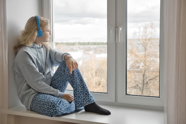 Una joven bonita se sienta en el alféizar de la ventana mira por la ventana y escucha música en los auriculares