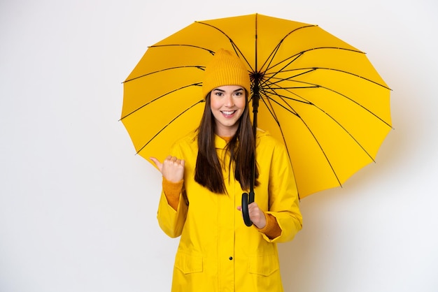 Joven brasileña con abrigo impermeable y paraguas aislada de fondo blanco apuntando hacia un lado para presentar un producto
