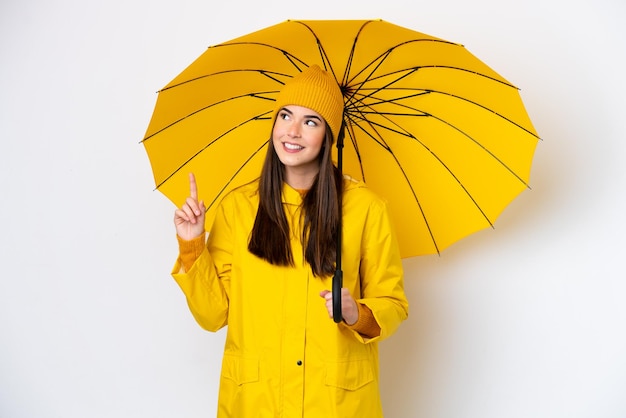 Joven brasileña con abrigo impermeable y paraguas aislado de fondo blanco señalando una gran idea