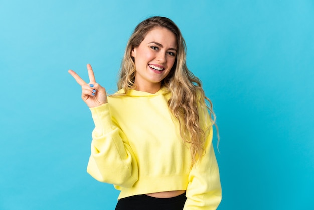 Joven brasileña aislada en azul sonriendo y mostrando el signo de la victoria