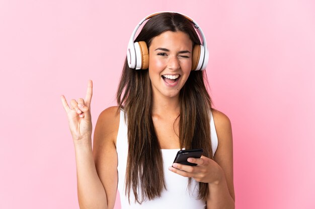 Joven brasileña aislada en la pared rosa escuchando música con un móvil haciendo gesto de rock