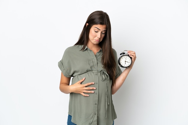Joven brasileña aislada sobre fondo blanco embarazada y sosteniendo el reloj con expresión estresada
