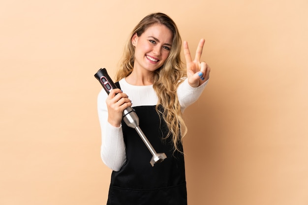 Joven brasileña con batidora de mano aislada en beige sonriendo y mostrando el signo de la victoria