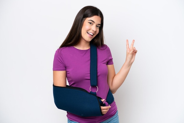 Foto joven brasileña con un brazo roto y usando un cabestrillo aislado de fondo blanco sonriendo y mostrando el signo de la victoria