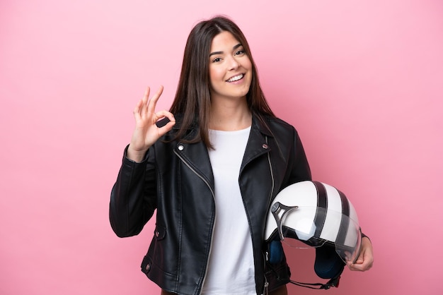 Joven brasileña con un casco de motocicleta aislado en un fondo rosado que muestra el signo de ok con los dedos