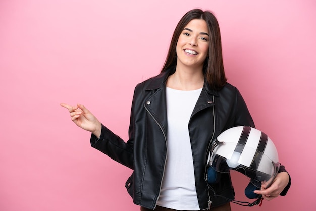 Joven brasileña con un casco de motocicleta aislado en un fondo rosado señalando con el dedo al costado