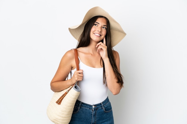 Joven brasileña con Pamela sosteniendo una bolsa de playa aislada sobre fondo blanco pensando en una idea mientras mira hacia arriba