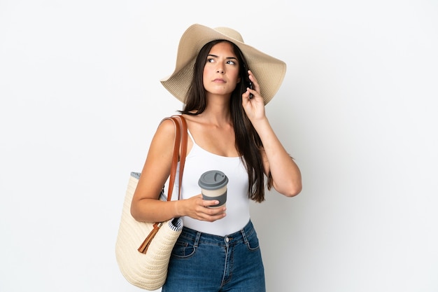 Foto joven brasileña con pamela sosteniendo una bolsa de playa aislada sobre fondo blanco sosteniendo un café para llevar y un móvil