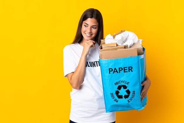 Joven brasileña sosteniendo una bolsa de reciclaje llena de papel para reciclar aislado sobre fondo amarillo mirando hacia el lado y sonriendo