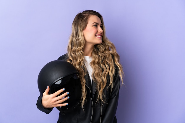 Foto joven brasileña sosteniendo un casco de moto aislado en púrpura mirando hacia un lado y sonriendo