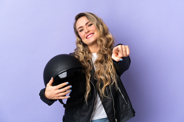 Joven brasileña sosteniendo un casco de motocicleta aislado en la pared púrpura apuntando hacia el frente con expresión feliz