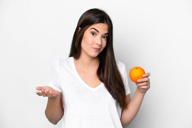 Joven brasileña sosteniendo una naranja aislada de fondo blanco haciendo gestos de duda mientras levanta los hombros