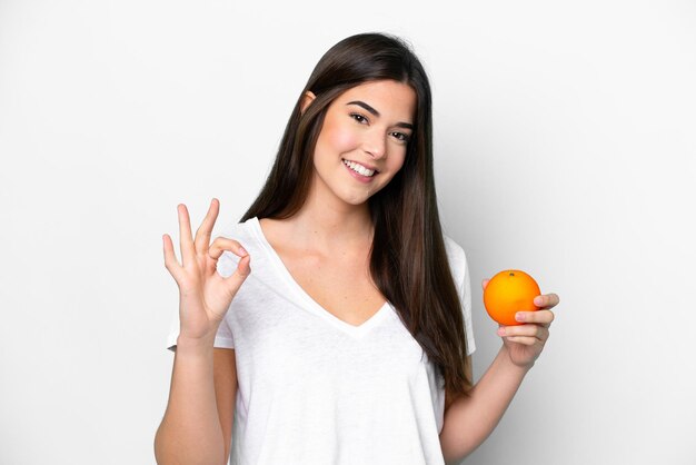 Joven brasileña sosteniendo una naranja aislada de fondo blanco mostrando el signo de ok con los dedos