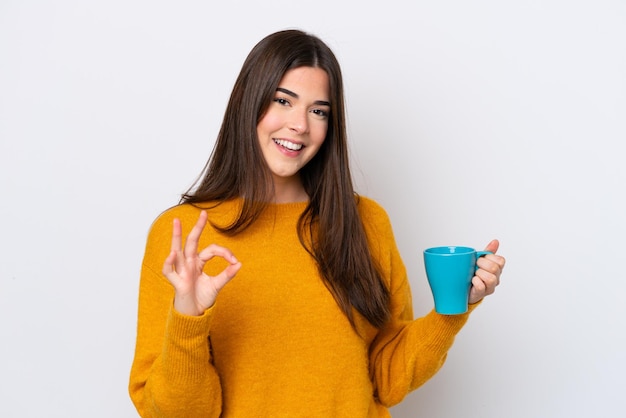 Joven brasileña sosteniendo una taza de café aislada de fondo blanco mostrando el signo de ok con los dedos