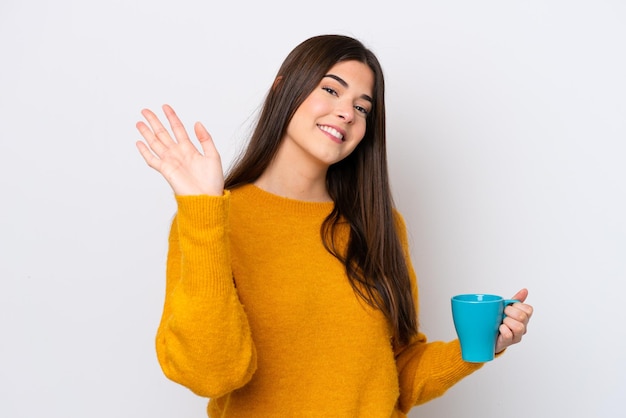 Joven brasileña sosteniendo una taza de café aislada de fondo blanco saludando con la mano con expresión feliz