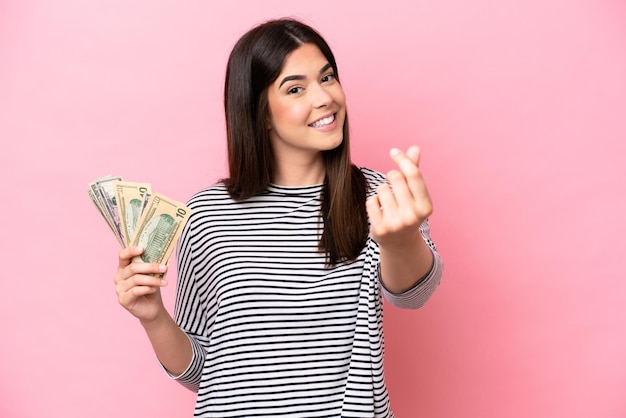 Foto joven brasileña tomando mucho dinero aislada de fondo rosa haciendo gestos de dinero