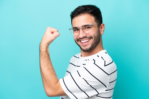 Joven brasileño aislado de fondo azul con gafas y celebrando una victoria