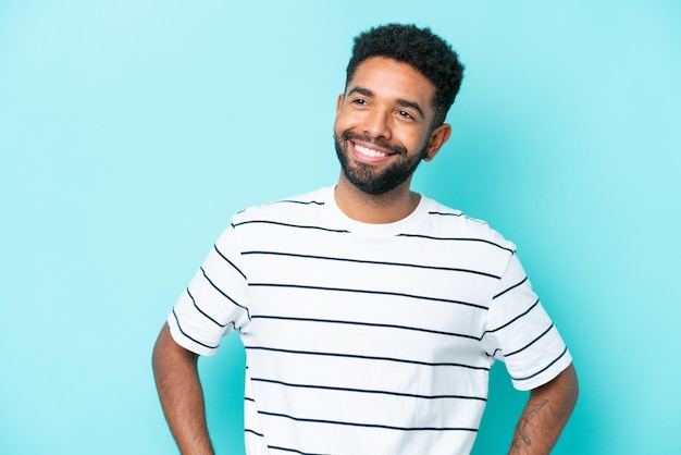 Joven brasileño aislado de fondo azul posando con los brazos en la cadera y sonriendo