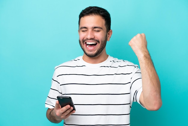 Joven brasileño aislado de fondo azul usando teléfono móvil y haciendo gesto de victoria