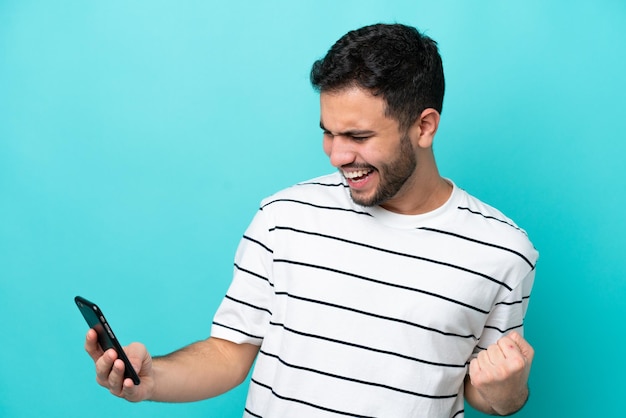 Joven brasileño aislado de fondo azul usando teléfono móvil y haciendo gesto de victoria