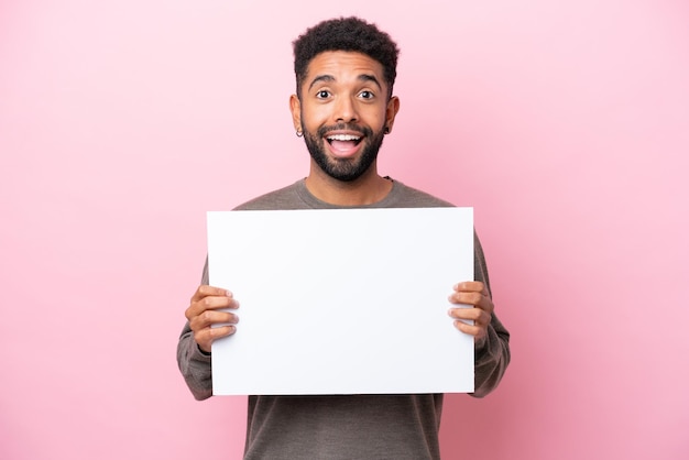 Joven brasileño aislado de fondo rosa sosteniendo un cartel vacío con expresión feliz