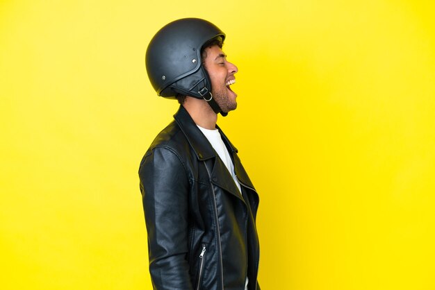 Joven brasileño con casco de motocicleta aislado de fondo amarillo riéndose en posición lateral