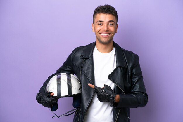 Joven brasileño con casco de motocicleta aislado de fondo morado apuntando hacia un lado para presentar un producto