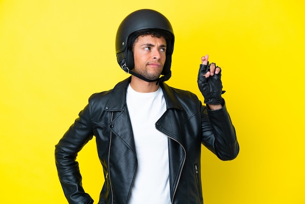 Joven brasileño con un casco de motocicleta aislado sobre fondo amarillo con los dedos cruzando y deseando lo mejor