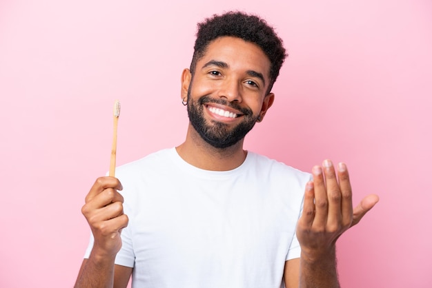 Joven brasileño cepillando dientes aislado en un fondo rosa invitando a venir con la mano Feliz de que hayas venido