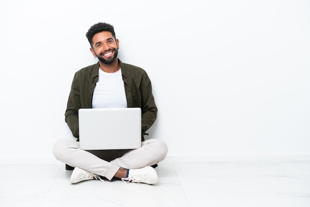 Joven brasileño con una computadora portátil sentada en el suelo aislada en blanco posando con los brazos en la cadera y sonriendo