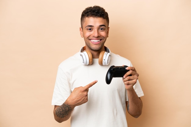 Foto joven brasileño jugando con un controlador de videojuegos aislado en un fondo beige apuntando hacia un lado para presentar un producto