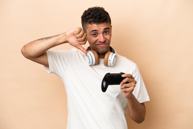 Joven brasileño jugando con un controlador de videojuegos aislado en un fondo beige mostrando el pulgar hacia abajo con expresión negativa