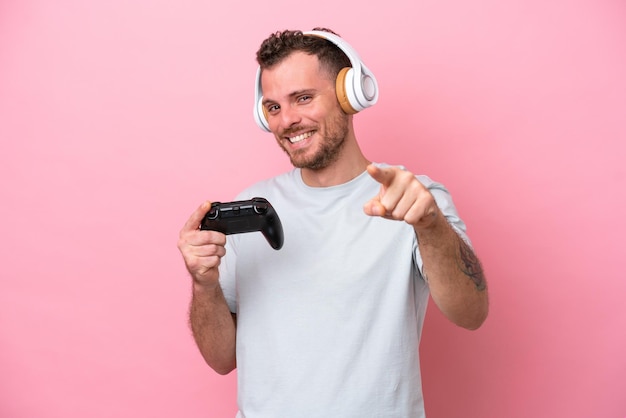 Joven brasileño jugando con un controlador de videojuegos aislado en un fondo rosa apuntando al frente con expresión feliz