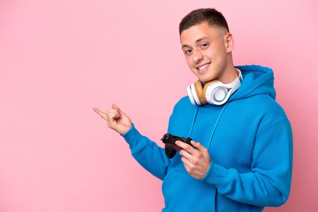 Joven brasileño jugando con un controlador de videojuegos aislado en un fondo rosa apuntando con el dedo hacia un lado