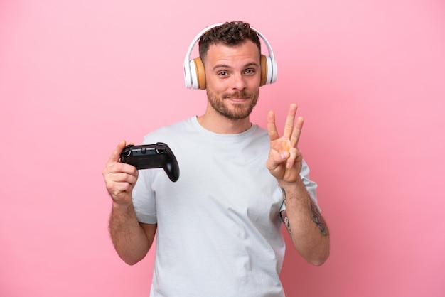 Joven brasileño jugando con un controlador de videojuegos aislado en un fondo rosa feliz y contando tres con los dedos