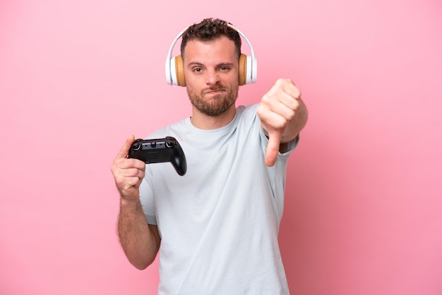 Joven brasileño jugando con un controlador de videojuegos aislado en un fondo rosa mostrando el pulgar hacia abajo con expresión negativa