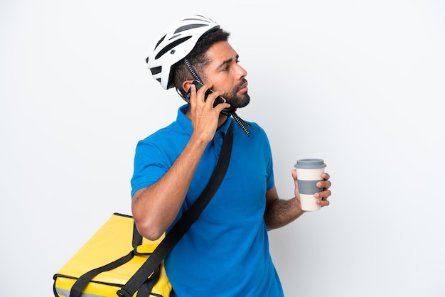 Joven brasileño con mochila térmica aislado de fondo blanco sosteniendo café para llevar y un móvil