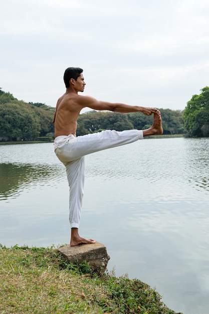 Joven brasileño de pie junto a un lago, sin camisa, estirando una de sus piernas.