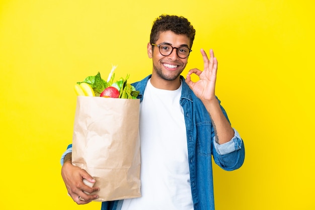 Joven brasileño sosteniendo una bolsa de compras aislada de fondo amarillo mostrando el signo de ok con los dedos