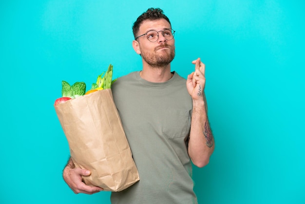 Joven brasileño sosteniendo una bolsa de compras aislada de fondo azul con los dedos cruzados y deseando lo mejor