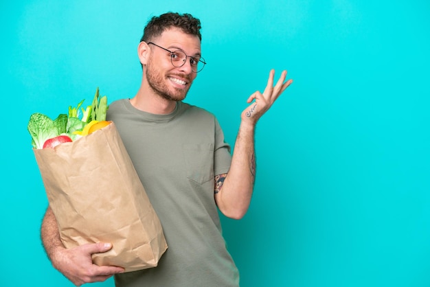 Joven brasileño sosteniendo una bolsa de compras aislada de fondo azul extendiendo las manos a un lado para invitar a venir