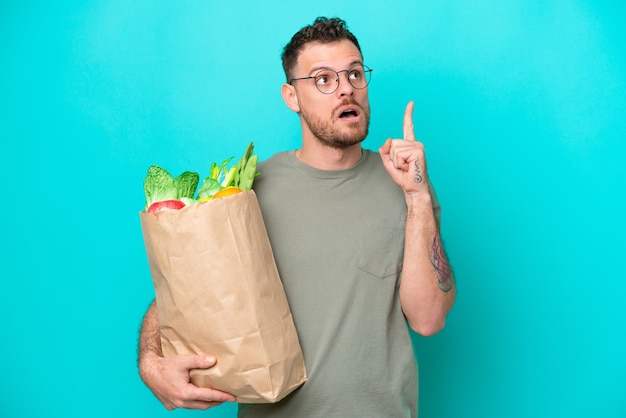 Joven brasileño sosteniendo una bolsa de compras aislada de fondo azul pensando en una idea apuntando con el dedo hacia arriba