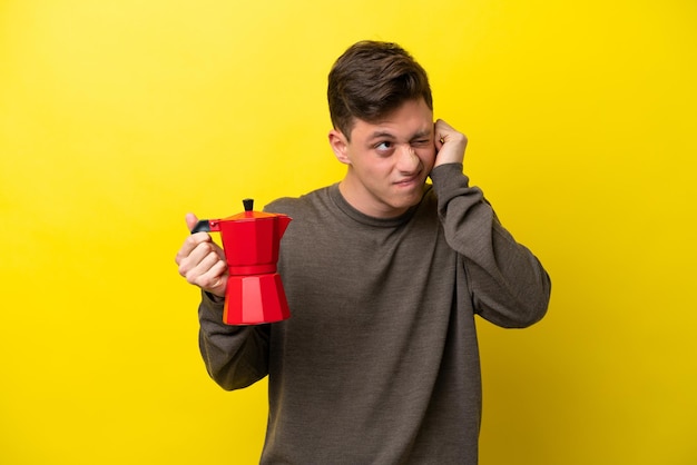 Joven brasileño sosteniendo una cafetera aislada de fondo amarillo frustrado y cubriendo las orejas