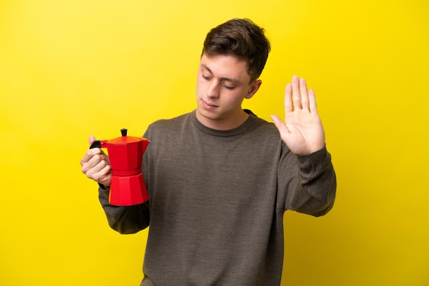 Joven brasileño sosteniendo una cafetera aislada de fondo amarillo haciendo un gesto de alto y decepcionado