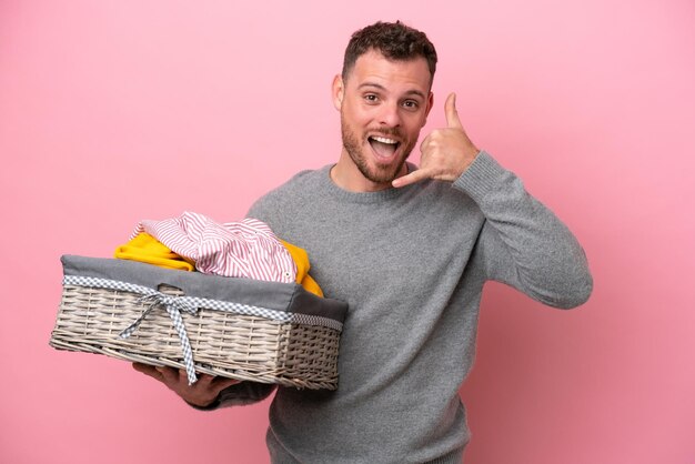 Joven brasileño sosteniendo una cesta de ropa aislada en un fondo rosa haciendo un gesto telefónico Llámame de nuevo