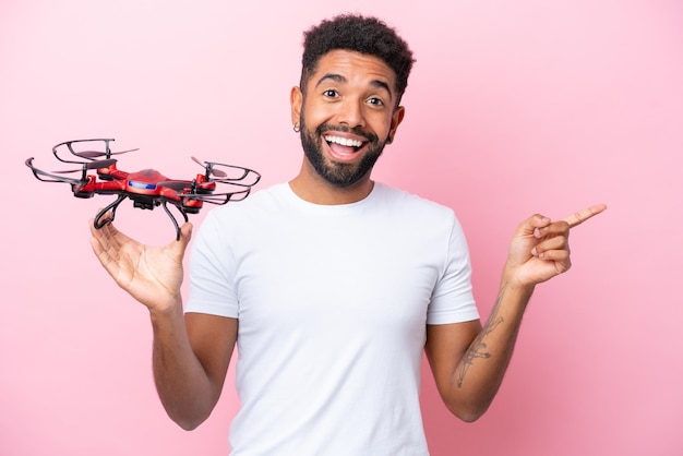 Joven brasileño sosteniendo un dron aislado de fondo rosa sorprendido y señalando con el dedo hacia un lado