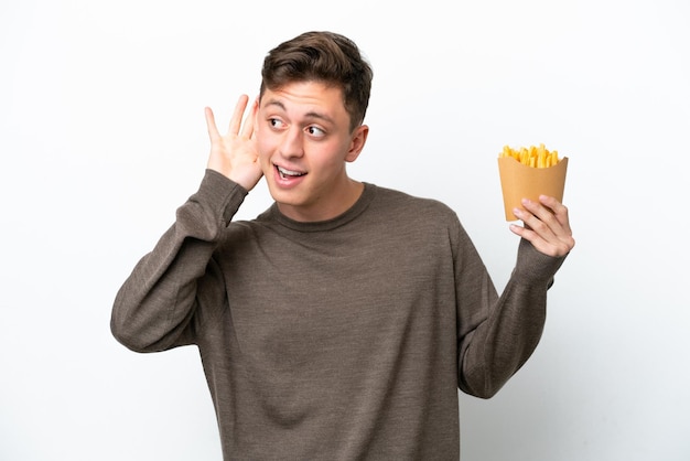 Joven brasileño sosteniendo patatas fritas aisladas de fondo blanco escuchando algo poniendo la mano en la oreja