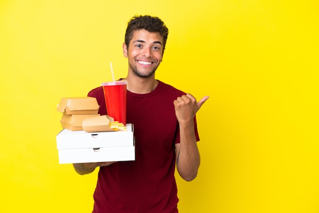 Joven brasileño sosteniendo pizzas y hamburguesas de fondo aislado apuntando hacia un lado para presentar un producto