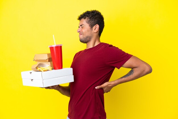 Joven brasileño sosteniendo pizzas y hamburguesas de fondo aislado que sufre de dolor de espalda por haber hecho un esfuerzo