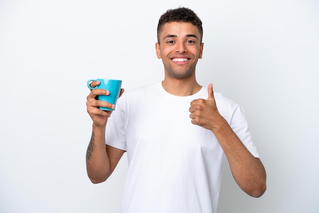 Joven brasileño sosteniendo una taza de café aislado de fondo blanco con los pulgares hacia arriba porque algo bueno sucedió